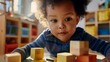 © iuricazac - Young child with curly hair wearing a blue striped shirt focused on wooden blocks in a brightly lit room with colorful shelves in the background.