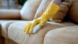 © iuricazac - Person wearing yellow gloves cleaning a beige couch with a white cloth.