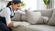 A woman in a cleaning uniform is meticulously dusting a beige sofa cushion.