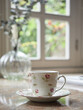 close-up view porcelain tea cup and saucer on kitchen countertop, background out of focus window and glass vase. Cozy cottage indoors.