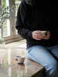 unrecognizable woman in jeans and black sweater sitting on the kitchen countertop next to the window holding a cup of hot tea.