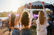 © artiemedvedev - Back view of three young women at beach music fest with raised drinks, enjoying concert vibes. Casual summer outfits, sunset lighting, crowd and stage in background, girls celebrate together outdoors.
