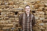 Fototapeta Panele - Confident Middle-aged Man in Checkered Shirt Posing in Front of Stone Wall