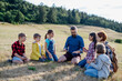 Children and teachers sitting on grass on meadow playing clapping game. Dedicated teachers during outdoor active education teaching about ecosystem, ecology.