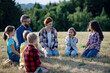 © Halfpoint - Children and teachers sitting on grass on meadow playing clapping game. Dedicated teachers during outdoor active education teaching about ecosystem, ecology.