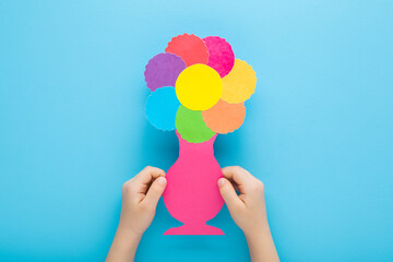 Wall Mural - Baby boy hands holding and showing beautiful flower head and vase shape from colorful paper on pastel blue table background. Closeup. Point of view shot. Little child creating gift. Top down view.