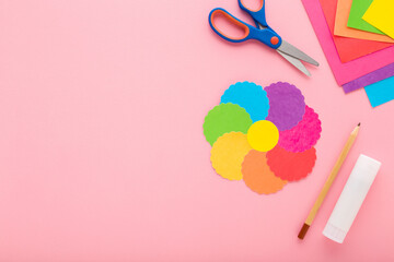 Wall Mural - Colorful flower shape, scissors, glue stick, pencil and application paper on light pink table background. Pastel color. Closeup. Making decoration elements. Empty place for text. Top down view.