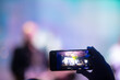 Person holding smartphone with background silhouettes of concert crowd with stage lights
