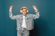 Mature female dancing with joy while wearing a denim outfit and headphones on a blue background