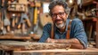 © Rattanathip - A handsome, bearded man in his 40s wearing glasses, a denim shirt, and suspenders is leaning on a wooden table in a workshop. He has a warm smile on his face.