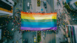 A rainbow flag is being carried by a crowd of people in a city. The flag is large and colorful, and the people are walking down the street. Scene is celebratory and joyful.
