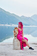 A woman with pink hair shares a lakeside moment with a Shiba Inu, the dog on a stone as they both look out over the water. The mountainous landscape reflects in the still lake