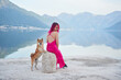 A woman with pink hair shares a lakeside moment with a Shiba Inu, the dog on a stone as they both look out over the water. The mountainous landscape reflects in the still lake