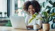 © venusvi - A smiling young woman sitting at a desk, working on a laptop, with a coffee mug and a plant beside her.