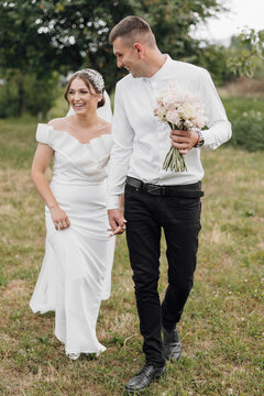 A bride and groom are walking together in a field, holding hands and smiling. The bride is wearing a white dress and the groom is wearing a white shirt and black pants