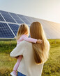 Rear view of mom holding her little girl in arms and showing at their house with installed solar panels. Alternative energy, saving resources and sustainable lifestyle concept. 