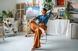 Satisfied dreaming artist woman resting on chair in art studio, smiling looks aside, pondering creative ideas for new pictures. Contented relaxed young female painter take break from painting process.