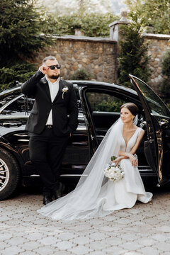 A man and woman are posing in front of a black car. The man is wearing a suit and sunglasses, while the woman is wearing a wedding dress. They are both smiling and seem happy