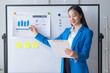 Asian businesswoman pointing at a white board with graphs on it. She is smiling and she is giving a presentation