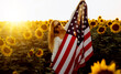 Beautiful young woman with the American flag in a sunflower field at sunset. 4th of July. Independence Day.
