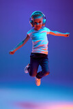 Fototapeta Sport - Schoolboy in casual attire jumping of joy while dancing during listening music in headphones in mixed neon light against blue background. Concept of music, dance, fashion and style, technology. Ad
