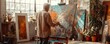© Влада Яковенко - Mature old man painting in messy and cluttered art studio with natural window light. The artist is surrounded by paint brushes and paints and is working on a canvas.