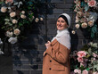 Portrait of a young caucasian woman dressed in a hijab near a wall with flowers.