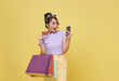 Excited Asian woman carrying shopping bags with credit card and looking mobile phone in hands isolated on yellow background.