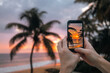 Close-up of hands holding smart phone. Man photographing sunset over ocean with mobile phone. Coconut palm trees on sand beach in south coast of Sri Lanka..