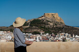 Fototapeta  - A woman in a hat and striped blouse standing in the grounds of San Fernando's Castle (Castell de Sant Ferran) looking towards Santa Barbara Castle of Alicante. Spanish holidays