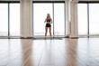 © sofiko14 - Fit young woman wearing workout gear stands on mat in spacious gym with large windows overlooking the city. Sports woman with dumbbells, ready for exercise routine, embodying health and fitness.