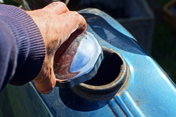 hand unscrews the round chrome shiny cap from a blue iron old dirty motorcycle tank during the day on the street