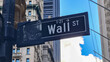 A close up on the sign, which points to the  Wall Street in New York City. There are tall buildings in the back. Famous landmarks. The sign has also a miniature of building along that street.