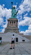 A man in shorts standing under an iconic representation of freedom and independence, the Statue of Liberty with flaming torch on Liberty Island. The Lady on a Pedestal is surrounded by clouds.
