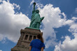 A woman in blue dress standing under an iconic representation of freedom and independence, the Statue of Liberty with flaming torch on Liberty Island. The Lady on a Pedestal is surrounded by clouds.