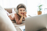 Fototapeta  - With headphones on, a sweet little girl, relaxing on a sofa, engrossed in the captivating content playing on her laptop