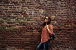 Beautiful brunette smiling woman in sunglasses looking up on the old red brick wall building in casual red shirt holding shoulder leather black bag on the spring city. Style banner