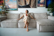 Smiling artist woman sitting on large sofa, relaxing. Businesswoman resting after work in new apartment, cozy interior, indoor plants, paintings. Happy girl in comfortable room, sweet home concept