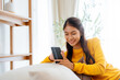 Happy young Asian woman sitting on sofa holding mobile phone using cellphone technology doing ecommerce shopping, buying online, texting messages relaxing on couch in cozy living room at home