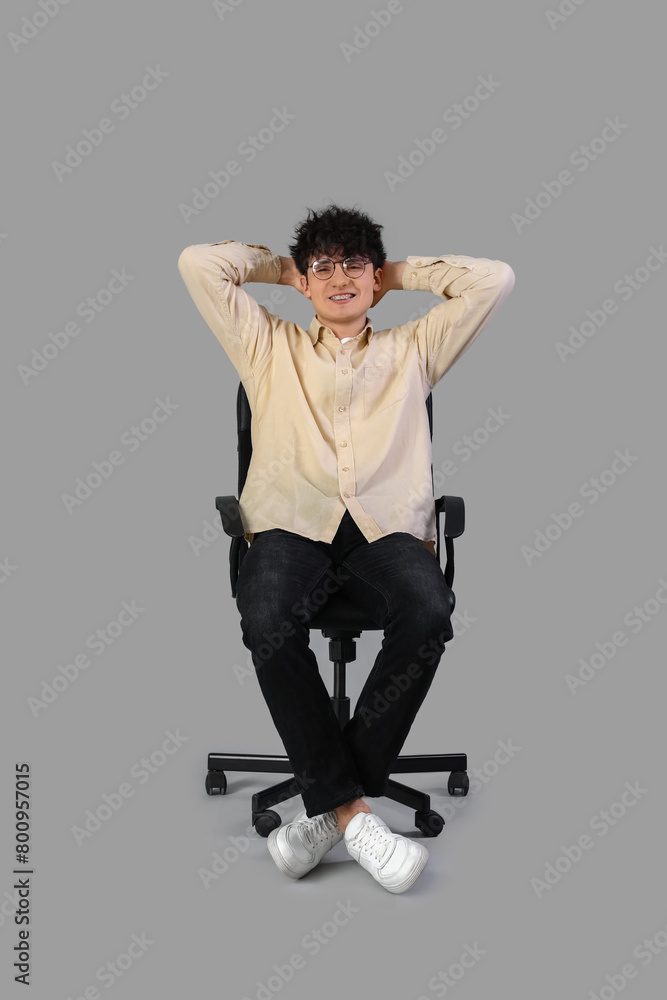 Young man in eyeglasses resting on office chair on grey background