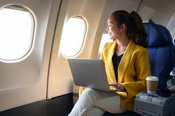 Confident businesswoman in formal clothes looking out of airplane window during flight while working on laptop computer Online internet connection, travel concept, business tourism.