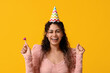 Beautiful young African-American woman in party hat with whistle on yellow background