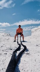 Wall Mural - Bodybuilder trains his muscles with cable on the hill under blue sky. Shirtless sportsman wearing cap and sunglasses doing workout with battle ropes in sunny summer day outdoors. Vertical video