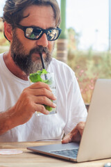 Wall Mural - Alternative office and workplace concept with young man drinking cocktail and using laptop on the cafe table in outdoor. Summer travel job digital lifestyle people. Handsome using computer connection