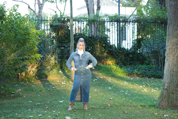 Pretty young blonde woman with a ponytail in her hair wearing a denim dress and brown boots posing in the park on the grass between the trees. The girl poses for photos. Beauty and fashion concept.