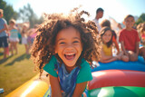 Fototapeta  - A young girl with curly hair is smiling and laughing while sitting on a bouncy castle. The scene is lively and fun, with other children around her, possibly playing or watching her