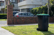 A household green garbage bin with wheels for garden waste is placed outside residential units on a grassy nature strip, awaiting collection. Copy space for your design.