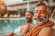 © yaqui_villegas - A joyful older man with a grey beard smiling warmly, wrapped in an orange towel by a swimming pool