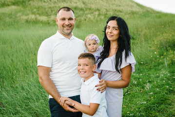 The son hugging parents in nature. Mom, dad, daughter girl and boy walk in the green grass in field. Happy young family spending time together outdoors. The concept of family holiday on summer day.