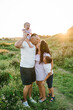 Mother kisses father, daughter girl and son boy stand in green grass in field. Children hugging parents in nature. Happy family spending time together outdoors. Family holiday at sunset on summer day